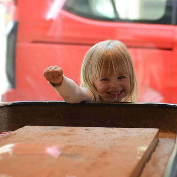 A young child smiling and raising a fist excitedly behind a glass case. The background features a red vehicle and blurred outdoor elements, suggesting an indoor play area near a parking lot. 