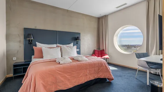Et moderne hotelværelse med en stor seng, et rundt vindue med udsigt over byen og et behageligt siddeområde.