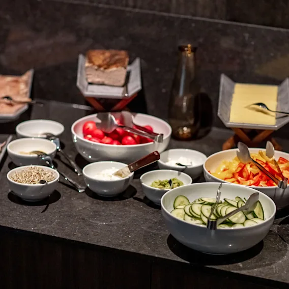 En mängd olika matvaror, inklusive skivade köttprodukter, ostar, grönsaker och röror, arrangerade på en mörk bänkskiva för en buffé.