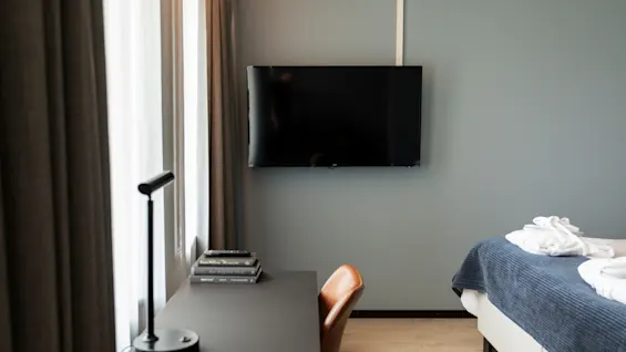 Et moderne hotellrom med skrivebord, stol, TV og en seng med blått sengetøy og hvite håndklær.