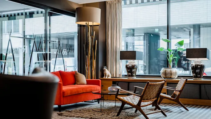 Rød sofa står i et moderne lobbyområde, omgitt av kurvstoler og et rundt bord. Store vinduer viser teksten "HOTEL", med en urban utsikt utenfor.