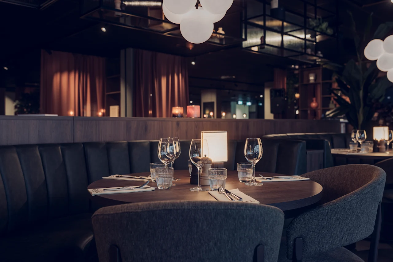 A round wooden table set with wine glasses, napkins, and candles sits in a dimly-lit restaurant with dark seating booths and soft overhead lighting. 