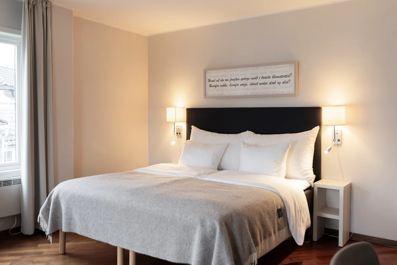 A comfortable hotel room with a bed, soft lighting, and a framed quote: Fuad vil du om frugten sponge midt i træets blomstertid?