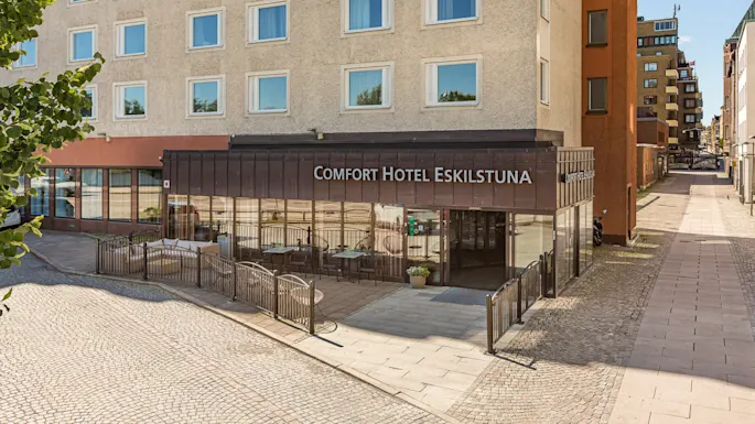 Comfort Hotel Eskilstunas inngang har uteservering med bord og stoler, plassert i et gatehjørne med brosteinsbelegning. Urban buildings surround the area.