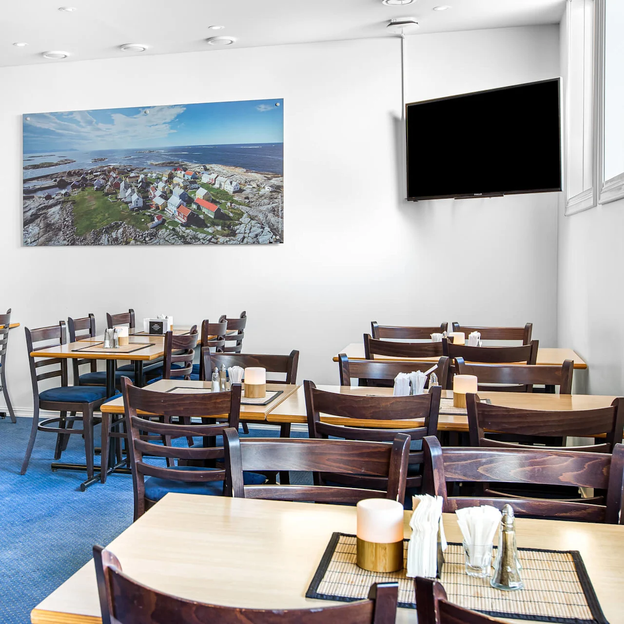 Restaurantbord og stoler er ordnet for servering, med et stort kystlandskapsmaleri på en hvit vegg og en svart TV-skjerm i nærheten. Gulvet er dekket med blått teppe.