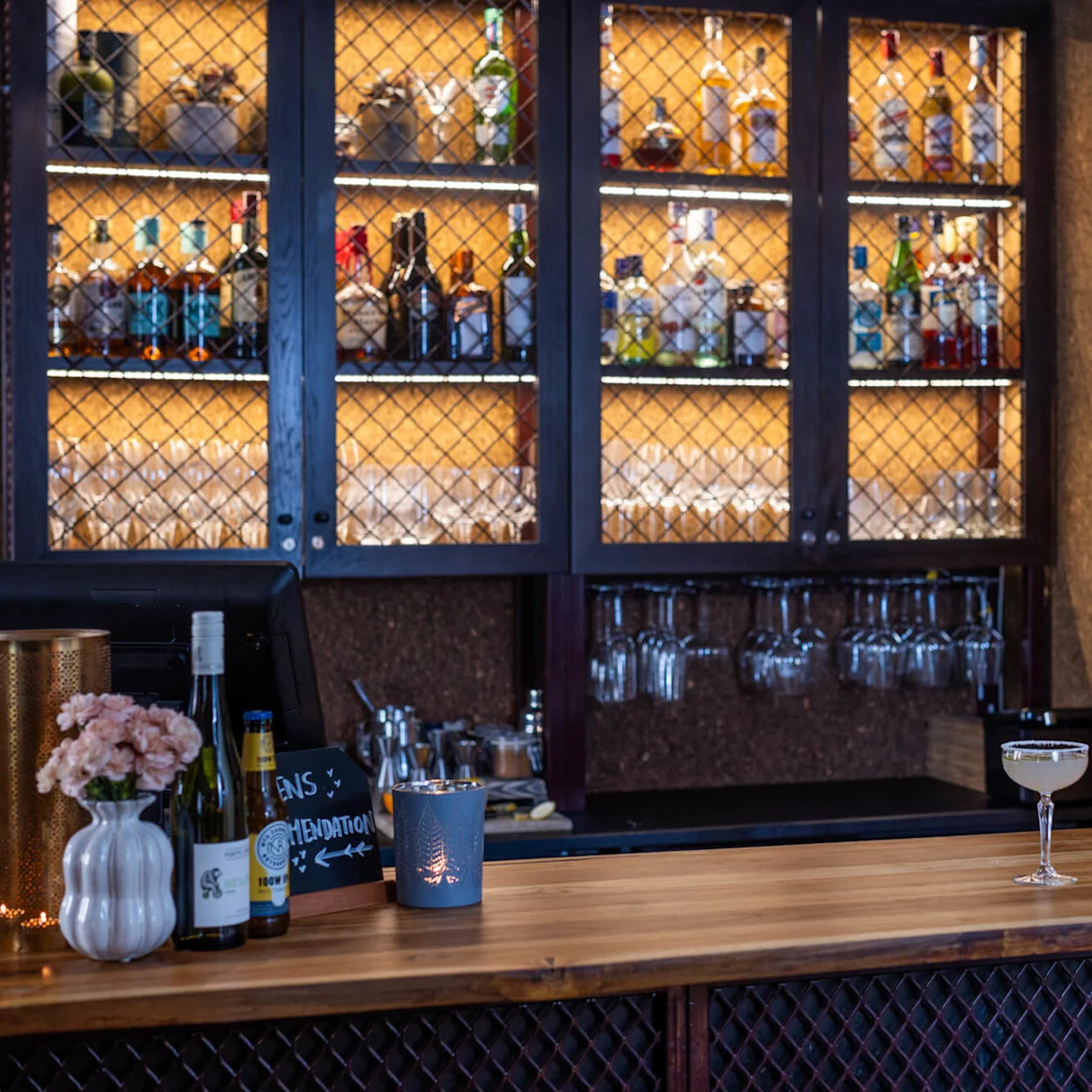 A cosy bar counter displays drinks and glasses with a chalkboard asking "IS IT DRINK O'CLOCK?"