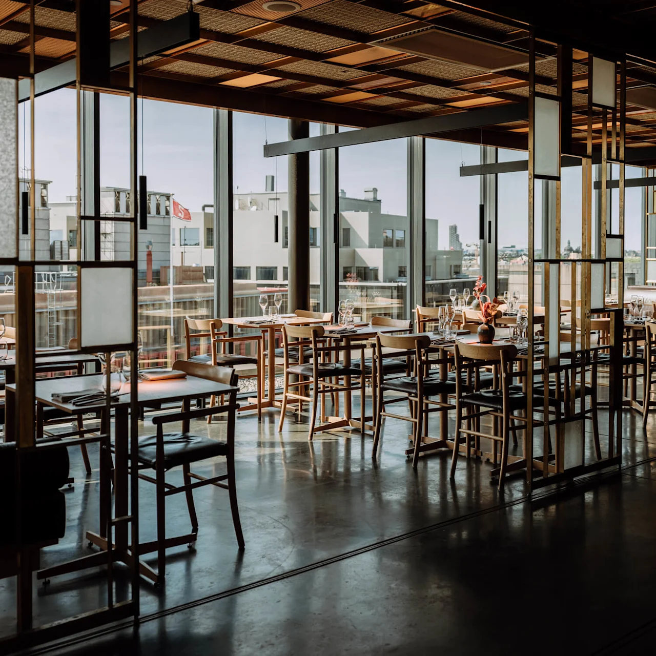 A dining area with wooden tables and chairs sits empty, surrounded by modern glass walls, overlooking a cityscape under clear blue skies.