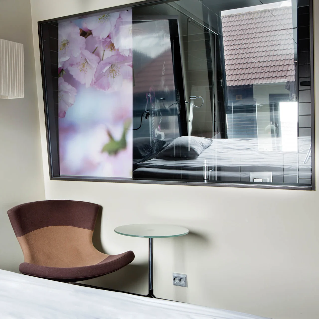En brun stol står ved siden av et glassbord i et moderne rom. Bak en stor blomsterkunstverk og et vindu som reflekterer en seng og ytre hustak.