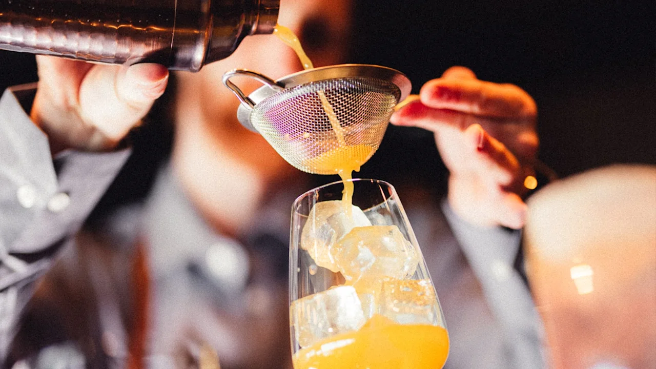 En bartender siler oransje væske fra en shaker i et glass fylt med isbiter, mot en uskarp innendørs bakgrunn.