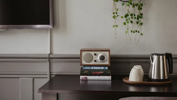 Ett hotellrumsbord med en radio, böcker inklusive UR SKOGENS DJUP, GROBYGGARNA, DANSARE, och en teservis, som erbjuder komfort och underhållning.