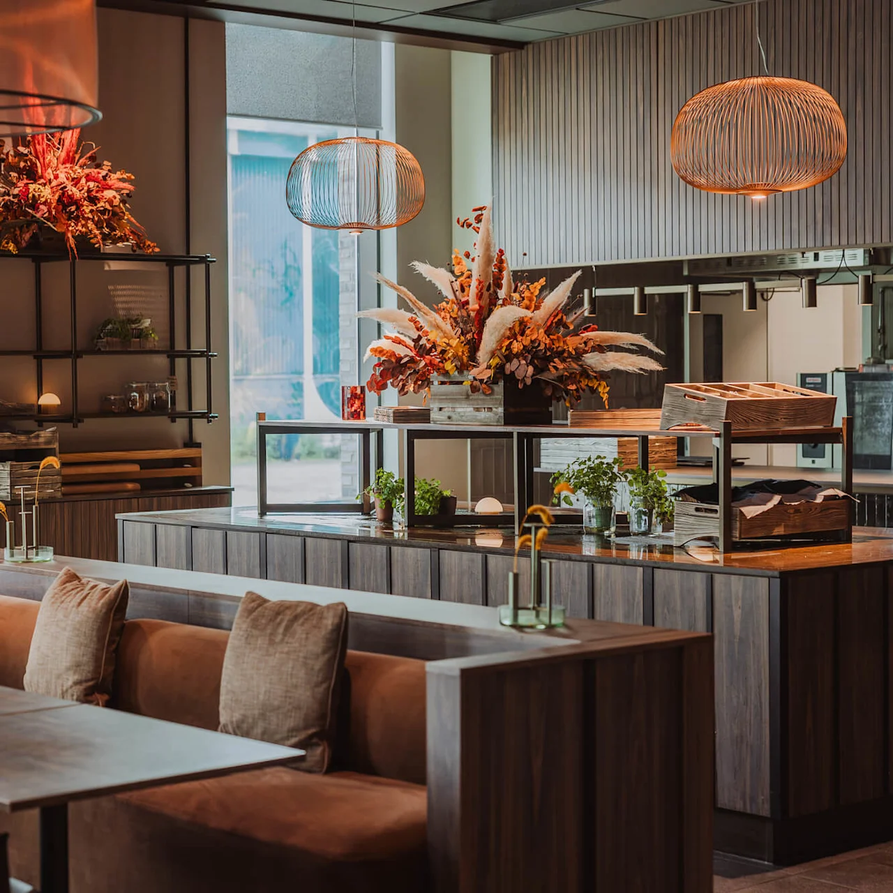 Modern café med träbord och bruna stoppade sittplatser. Dekorativa blomsterarrangemang står på bänkar under varma pendellampor. Naturligt ljus tränger in genom stora fönster och skapar en mysig atmosfär.