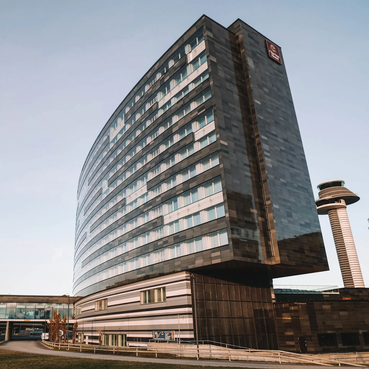 A tall, modern hotel building with a glass facade stands beside an airport control tower, under a clear sky. A skywalk connects it to adjacent structures.