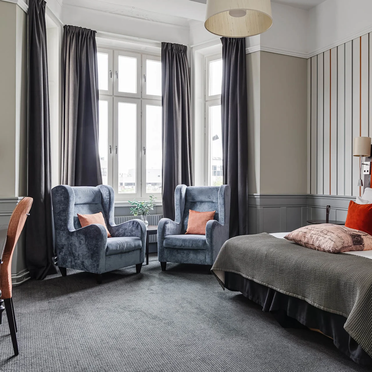 A stylish bedroom with a red headboard, two blue armchairs, and a large window with dark curtains.