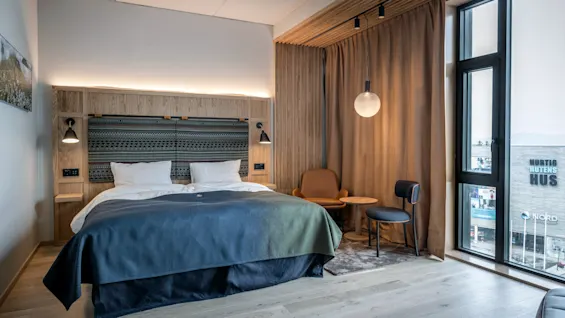 En pænt redt seng med et blåt tæppe og to puder står i et moderne rum. Til siden en siddegruppe med stole og et lille bord under en hængende lampe. Udenfor er en bygning synlig med tekst: "HURTIG RUTENS HUS" og "NORD".