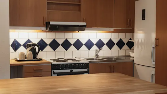 Et hyggeligt køkken med træskabe, flisebelagt stænkplade, komfur og vask. En elkedel og kopper står på bordpladen.