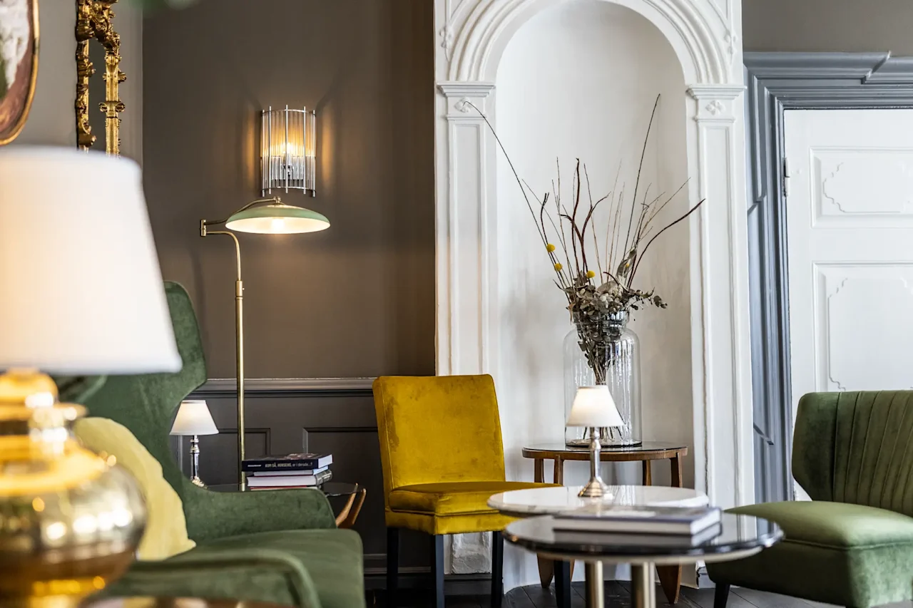 En hyggelig hotellounge med grønne og gule stole, lamper og et sofabord, der skaber en indbydende atmosfære.