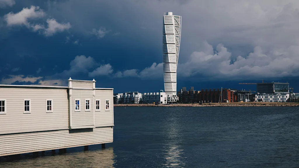 A view of Malmö's waterfront with a modern twisted skyscraper and a traditional building over the water under a cloudy sky.