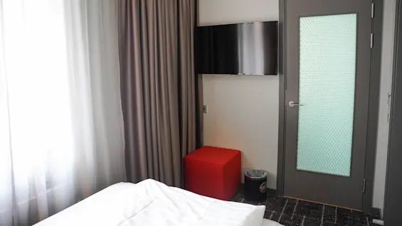 Et hotellromhjørne med en seng, flatskjerm-TV på veggen, gardiner for et vindu, og en lukket dør med frostet glass.