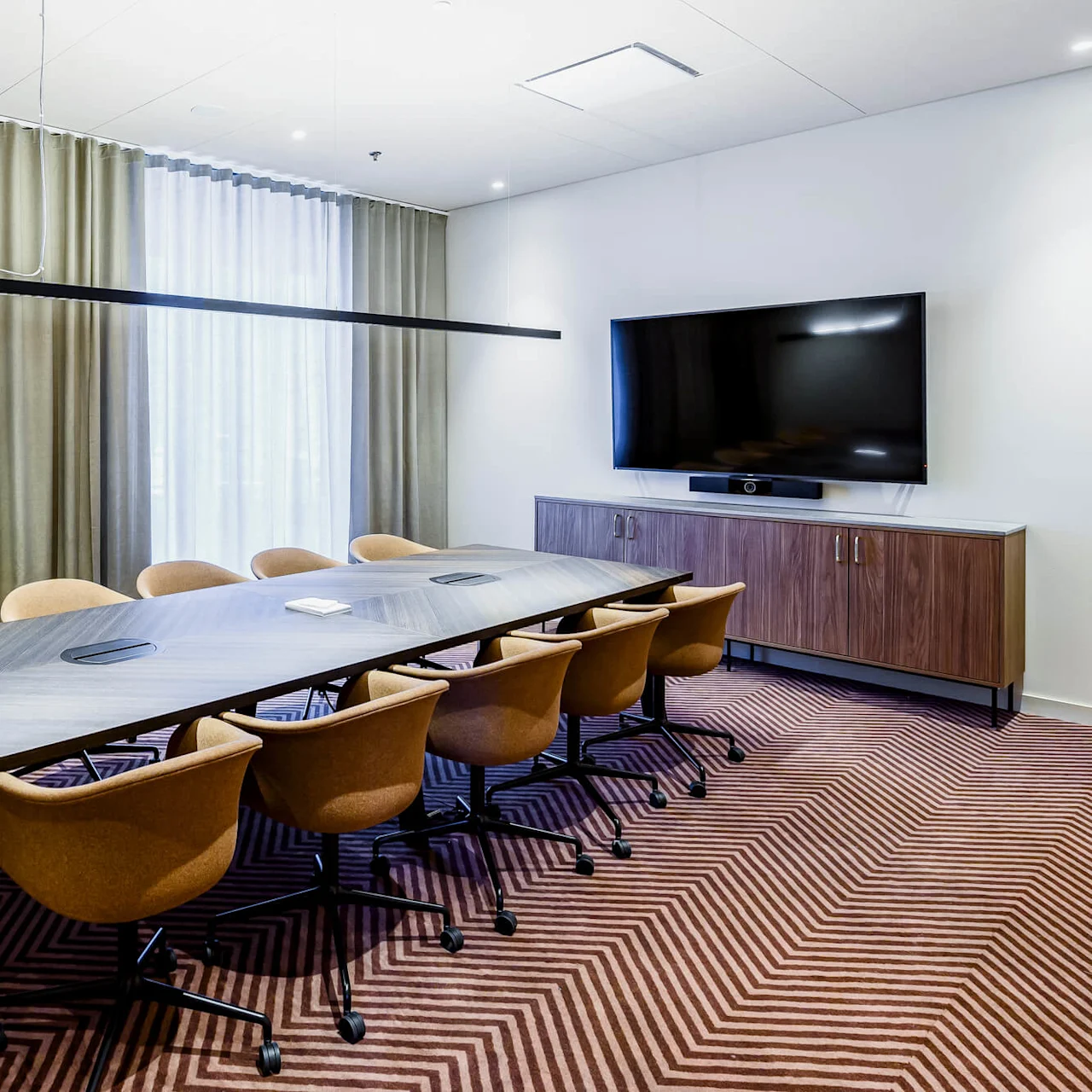 A long conference table surrounded by ten chairs rests on a patterned carpet. A large TV is mounted on the wall, situated above a wooden cabinet. Neutral-toned curtains cover the windows.