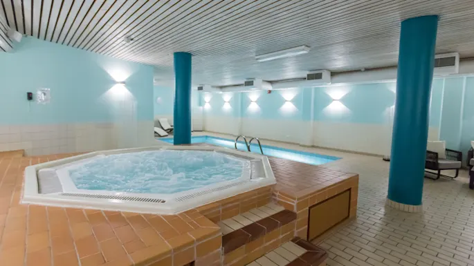 Et boblebad bobler i forgrunnen, omgitt av mursteinsfliser. I bakgrunnen ses et innendørs svømmebasseng i et lett opplyst rom med lyseblå vegger.