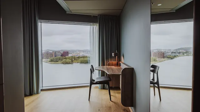 En stol står ved siden af et skrivebord af træ vendt mod et stort vindue med udsigt over byens skyline og flod, omgivet af vægge i neutrale farver og lyst trægulv.