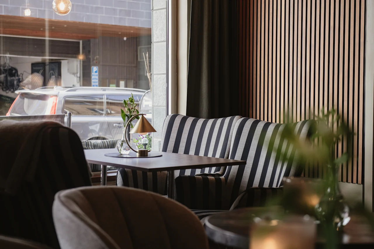 Mysig kafé sittgrupp med randig soffa och ett litet bord med lampa och vas. Stort fönster visar en parkerad bil utanför. Neutral, stilren inredning.