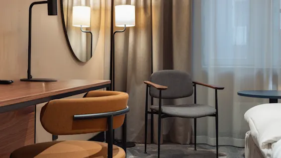 Et moderne hotelværelse med et skrivebord, lampe og stol ved siden af et rundt spejl. Gardiner indrammer et vindue, og en anden stol står ved siden af sengen.