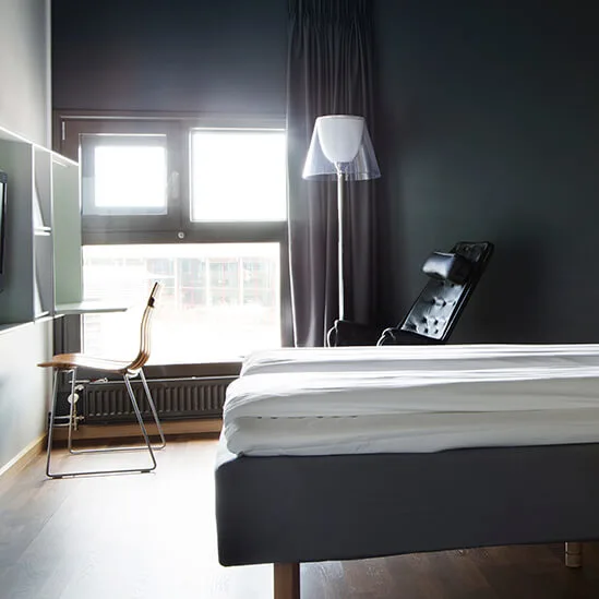 Moderni makuuhuoneessa on sänky, jossa on valkoiset vuodevaatteet tummalla seinällä. Vasemmalla ikkuna valaisee tuolin ja työpöydän. Huoneessa on minimalistinen sisustus.