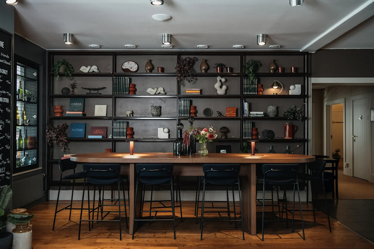 En stilfull lounge med ett långt gemensamt bord och barstolar, med en stor bokhylla fylld med inredning. Synlig text: WINE, CHESS, SOLITAIRE.