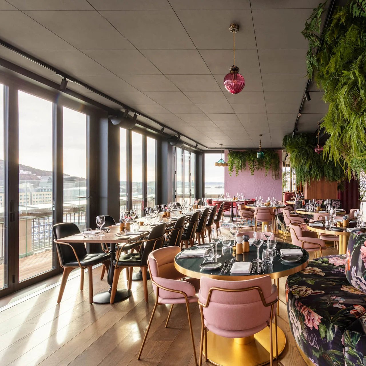 Restaurant tables arranged under hanging greenery, with upholstered chairs and floral-patterned booths. Floor-to-ceiling windows provide city views, creating an elegant dining atmosphere.