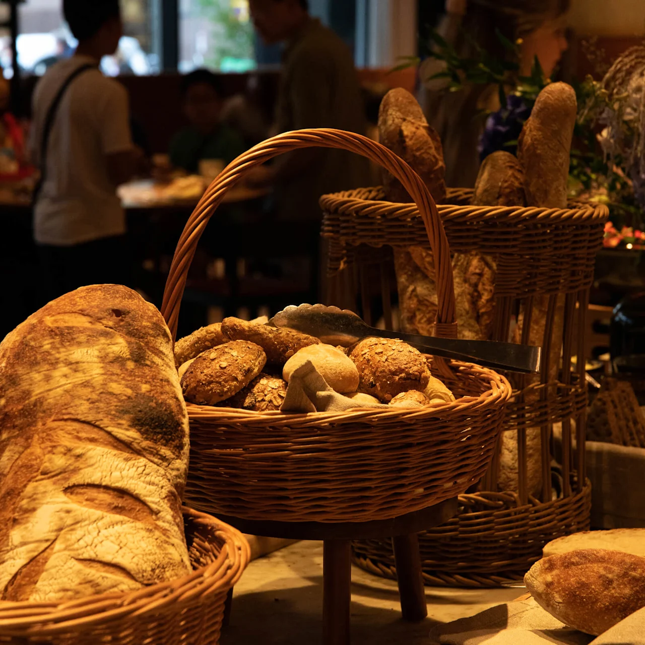 Baskets filled with various breads are placed on a marble countertop. In the background, people are gathered in a warmly lit room, creating a cozy atmosphere.