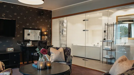Ylellinen hotellihuone, jossa on viihtyisä oleskelutila ja moderni kylpyhuone lasiseinän takana.