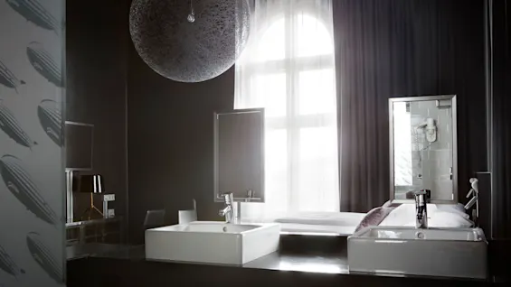 A modern bathroom with two rectangular sinks sits on a dark counter. A large decorative ceiling sphere hangs. A window with sheer curtains provides soft light.