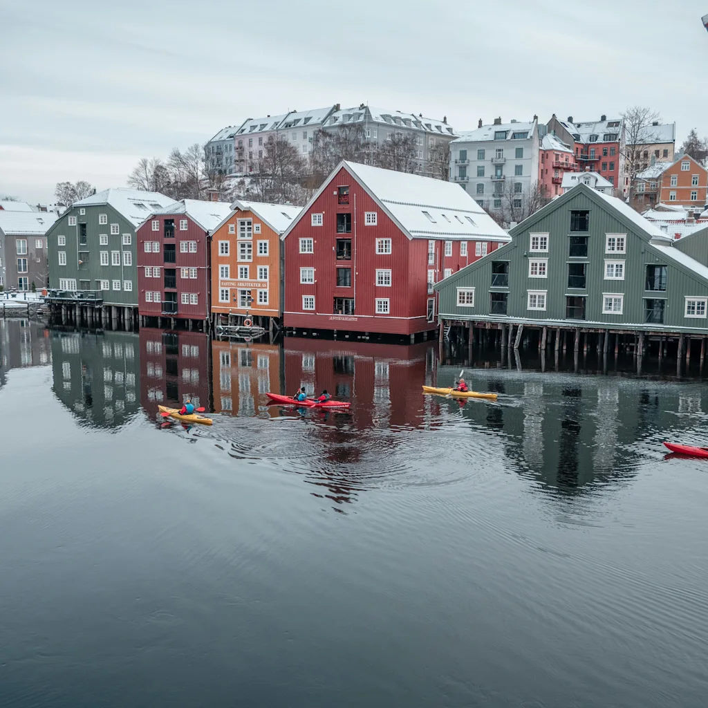 Kajakroere padler forbi farverige, snedækkede bygninger spejlet i roligt vand under en vinterhimmel.