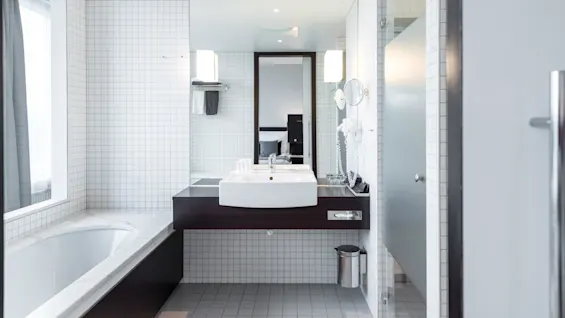 Et moderne bad har et hvitt badekar og servant, reflektert i store speil. Flisbelagte vegger og gulv skaper en ren, minimalistisk atmosfære med taklys som forsterker lysstyrken.