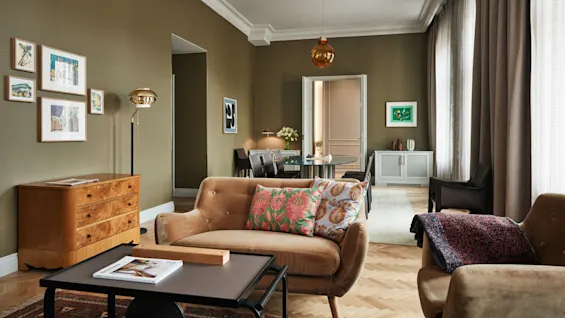 Et koselig stue med en beige sofa med fargerike puter står i et romslig, varmt opplyst område. Kunst preger olivenfargede vegger, med et treskap og elegant dekor.