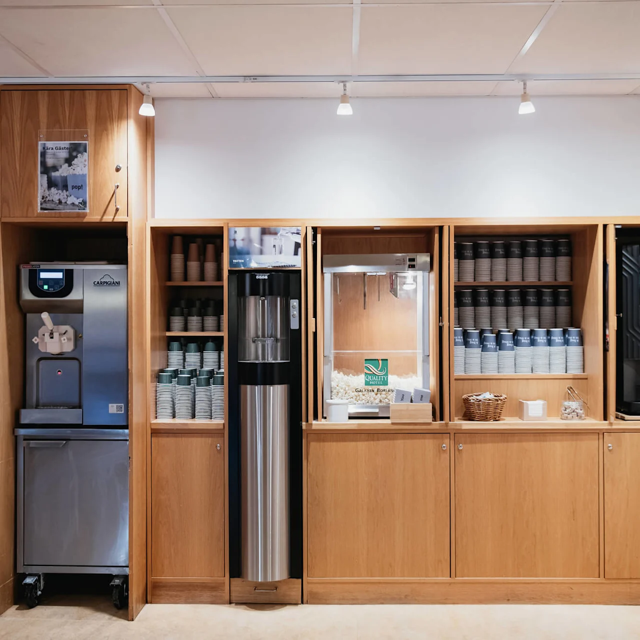 Soft serve machine stands in a wooden cabinet beside cups and a popcorn machine. Context: café environment. Text: "Göran Claesons Vingelvagn" and "pop!"