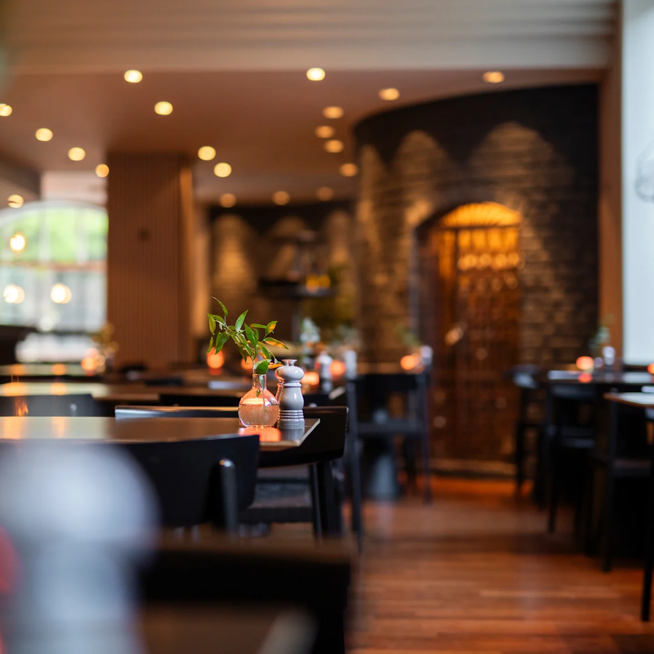 Borde med små vaser indeholder friske blomster, arrangeret i en svagt oplyst restaurant med trægulve, sten vægge og blød belysning, hvilket skaber en indbydende, hyggelig atmosfære.