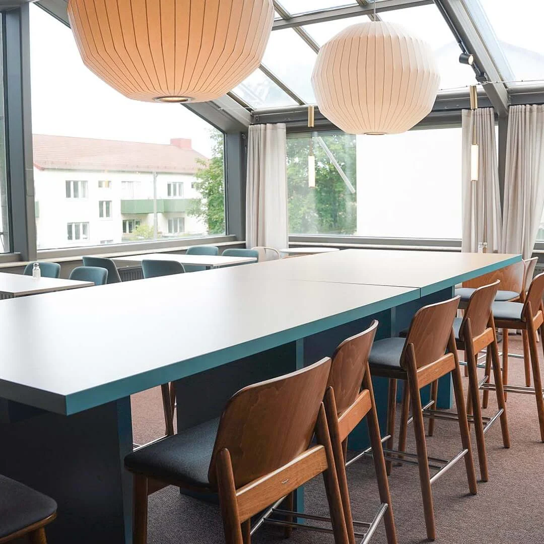 A spacious room with a long table, chairs, pendant lights, and large windows overlooking apartment buildings.