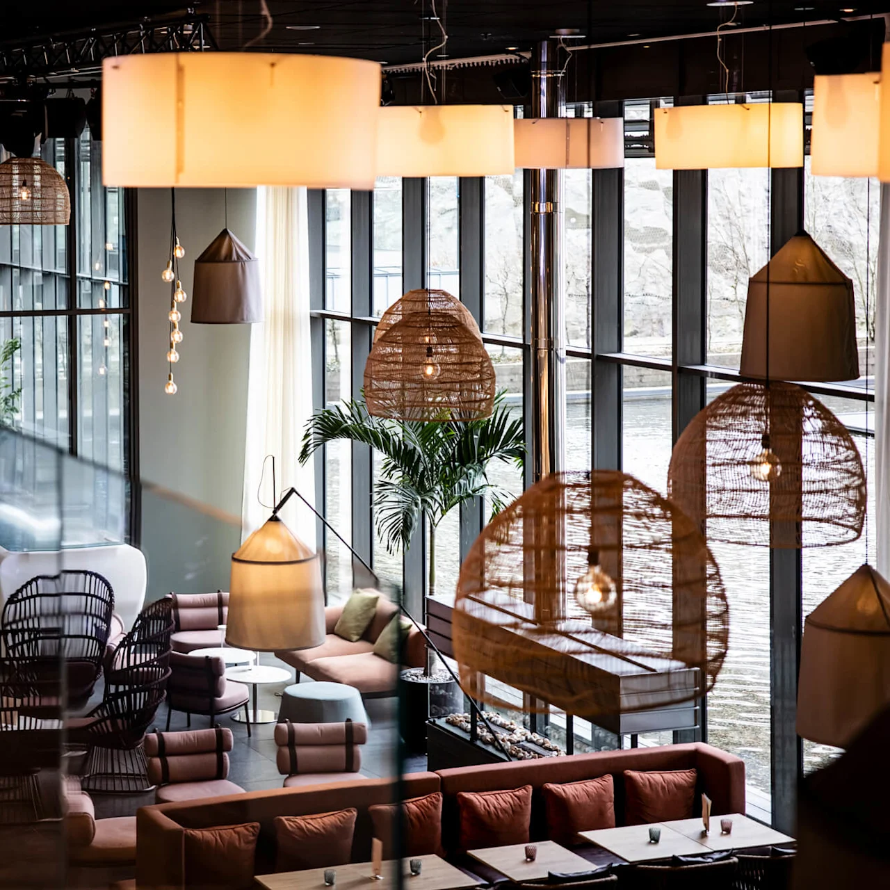 Luksuriøst loungeområde med kurvstole, loftslamper og en diskokugle; mod store vinduer ud til vandet. Indendørsplanter tilføjer grønne elementer til de elegante omgivelser.