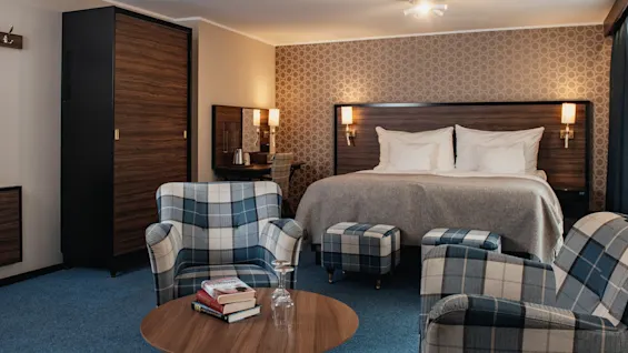 Viihtyisä hotellihuone, jossa on iso sänky, kaksi ruudullista nojatuolia, sohvapöytä kirjoineen ja työpöytäalue.