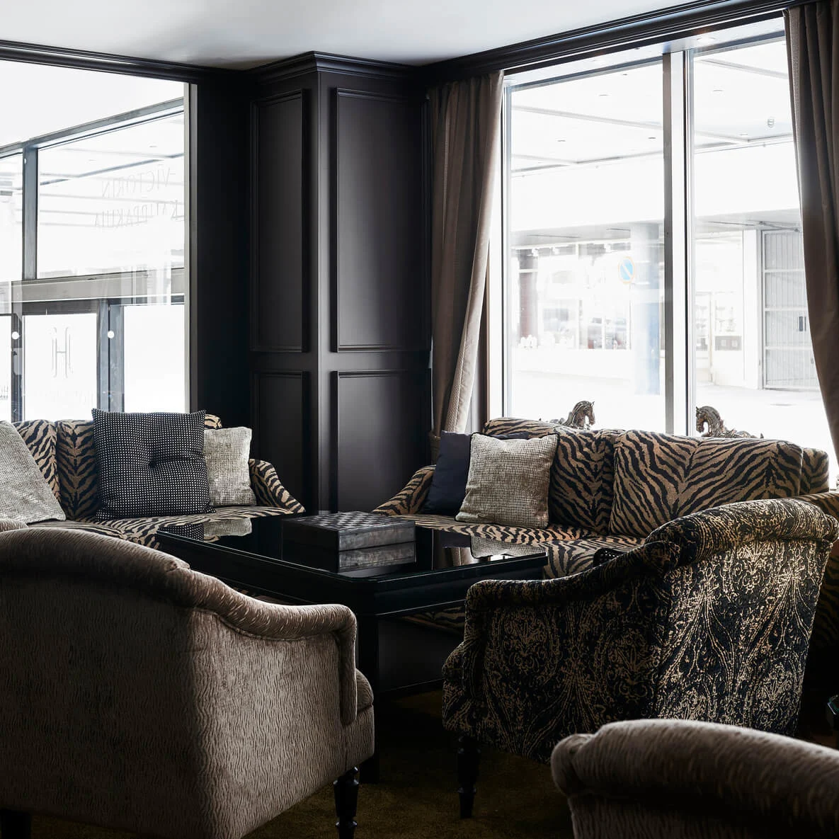Elegant lounge area features patterned sofas and armchairs centered around a glossy black table. Soft lighting includes a lamp on a side table near large windows.