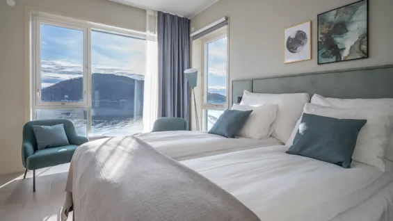 Et lyst hotelværelse med to senge, et hyggeligt siddeområde og et stort vindue med udsigt over en sø og bjerge.
