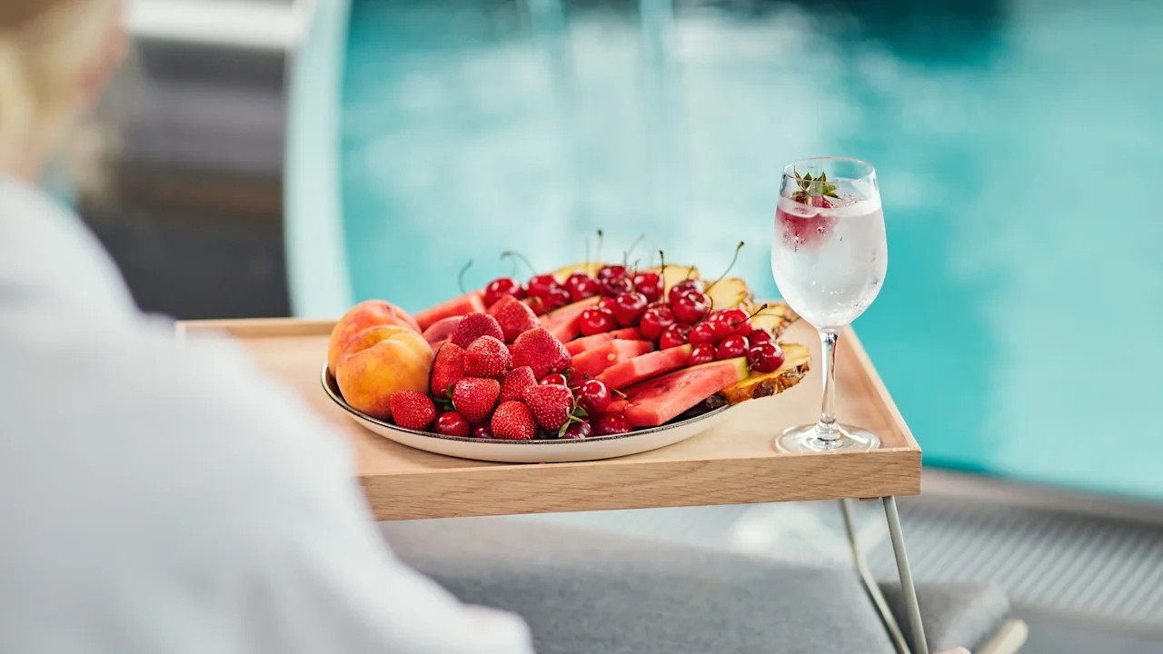 A tray filled with assorted fruits like watermelon, strawberries, and cherries, accompanied by a glass of iced water, is placed poolside on a wooden table.