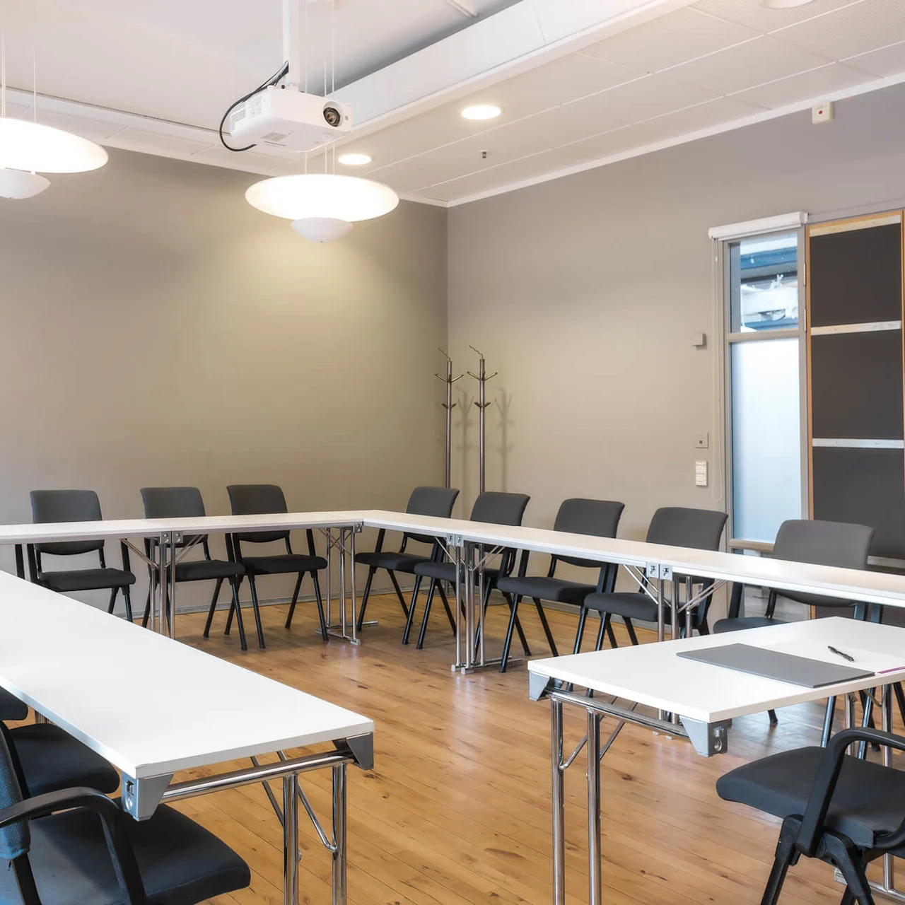 Conference room with U-shaped table arrangement, surrounded by black chairs, under ceiling lights. A projector hangs from the ceiling, and wooden flooring completes the modern, minimalist setting.