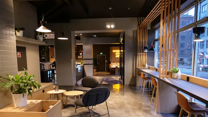 A modern cafe interior with wooden furniture and potted plants. High chairs line the window bar, and cushioned seating surrounds small round tables. The sign reads "WORK CHILL HERE."