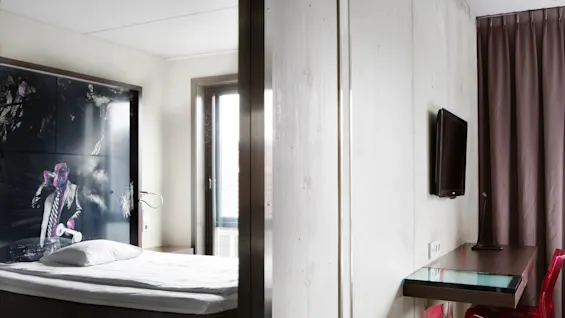 Seng med pute ved siden av kunstverk som viser mann, moderne hotellrom. Glasskrivebord med TV montert på vegg; rød stol; stort vindu med gardin i nærheten.