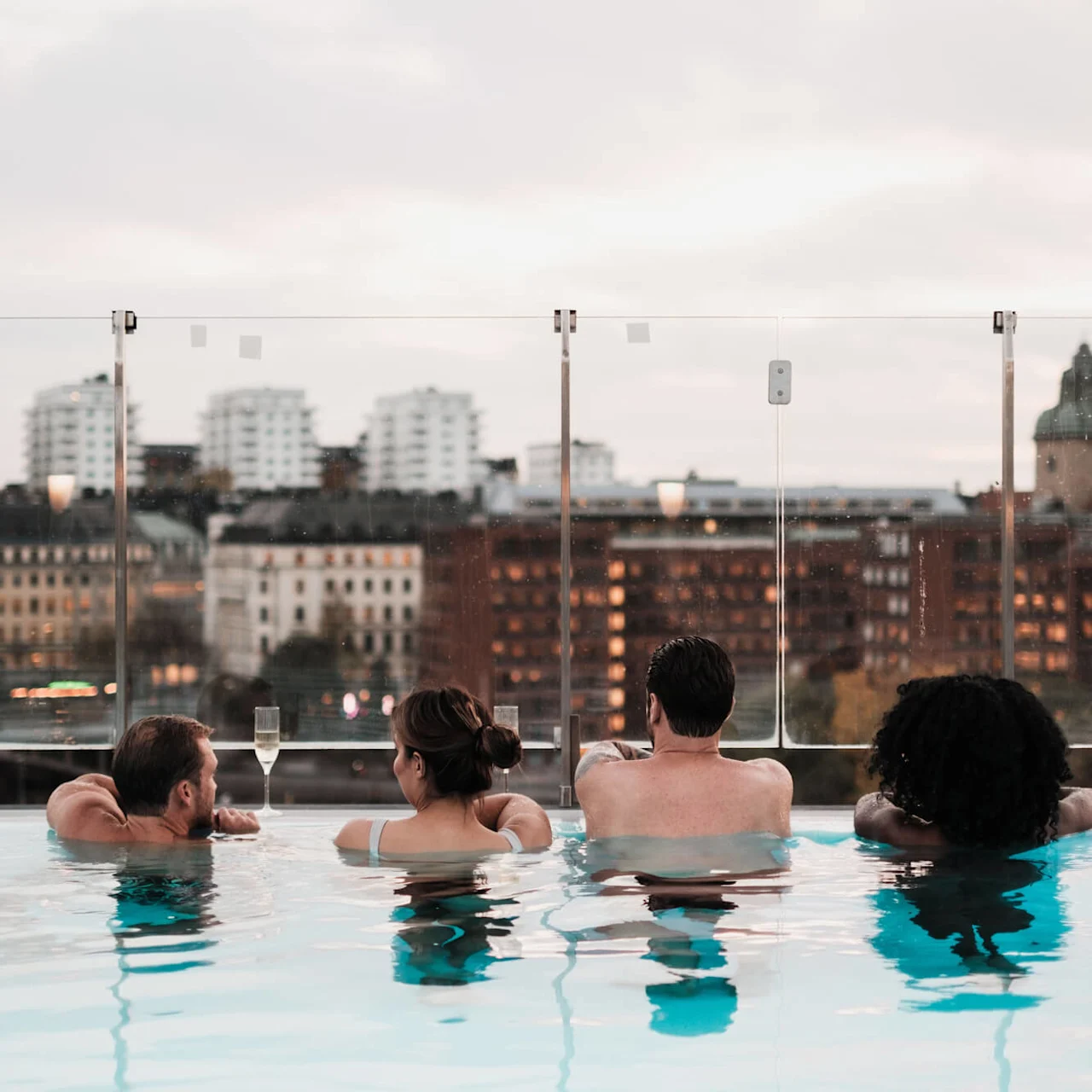 Fyra personer kopplar av i en takpool med utsikt över en stadsmiljö av byggnader och glas. Text: "#POOLCLUB @SELMACTYSPASTHLM".
