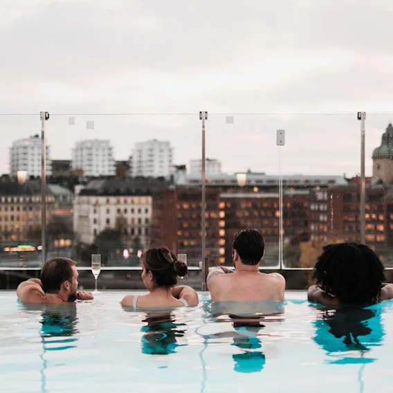 Fyra personer kopplar av i en takpool med utsikt över en stadsmiljö av byggnader och glas. Text: "#POOLCLUB @SELMACTYSPASTHLM".
