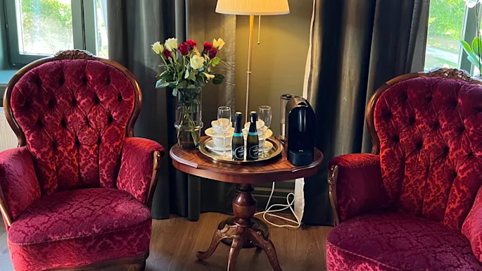 To utsmykkede røde lenestoler flankerer et rundt trebord med kaffemaskin, kopper og flasker, i et koselig rom med en stålampe, blomster og innrammet kunst.